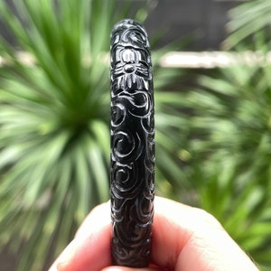 May include: A black, carved jade bangle bracelet. The bracelet features a detailed floral and swirling pattern. The dark green jade is polished and reflects light. The bracelet is held in a hand against a blurred green background.