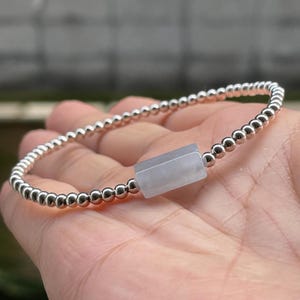 May include: A stretch bracelet featuring a series of small, polished, reflective silver-toned spherical beads arranged in a continuous loop, centered with a single, smooth, rectangular, pale lavender-gray stone bead.