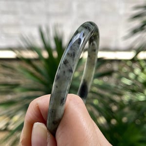 Op de afbeelding: Een gladde, gepolijste armband met een doorschijnende, lichtgrijze basis met donkergrijze en zwarte gevlekte insluitingen door de hele steen. De cirkelvormige band heeft een afgerond profiel en een glanzende afwerking die licht reflecteert.