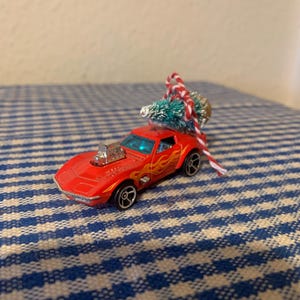 Puede incluir: Un coche de juguete rojo con llamas pintadas en el lateral lleva un pequeño árbol de Navidad artificial. El coche está sobre una superficie a cuadros azules y blancos.
