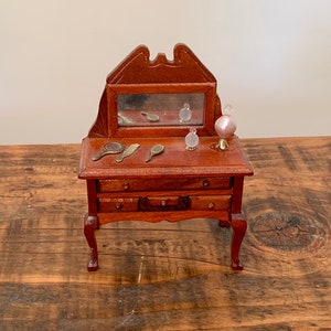Könnte beinhalten: Ein Miniatur-Schminktisch aus Holz mit einem Spiegel, drei Schubladen und zwei kleinen Flaschen. Der Schminktisch ist braun und hat ein geschnitztes Design auf der Oberseite. Er steht auf einer Holzoberfläche.
