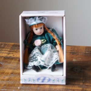 May include: A porcelain doll in a green dress with white lace trim and a pink flower. The doll has long red hair and is wearing a white hat. The doll is packaged in a white box with a blue logo.