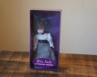 May include: A purple box with a porcelain doll inside. The doll is wearing a white blouse with a black and white plaid skirt. The box says "Miss Julie a Victorian teacher".
