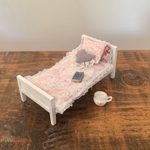 May include: A miniature white wooden bed with a pink and white lace bedspread, a purple heart-shaped pillow, and a small book. A white teacup with floral designs sits on the wooden surface.