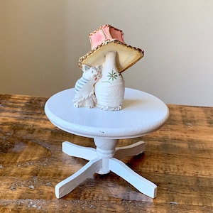 Puede incluir: Una figurita de cerámica blanca de un gato con un sombrero de pantalla de lámpara rosa y dorado sobre una mesa de pedestal blanca.