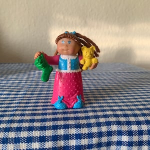 Op de afbeelding: Een Cabbage Patch Kid pop met bruin haar, die een roze jurk met witte stippen, een blauw vest met hartjes en blauwe konijnenpantoffels draagt. De pop houdt een groene sok en een gele teddybeer vast.