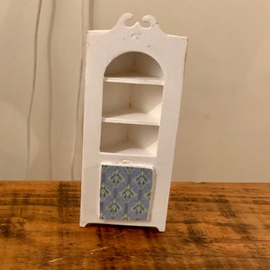 Op de afbeelding: Een wit hoekkastje met drie planken en een deur met een patroon. Het kastje is gemaakt van hout en heeft een eenvoudig ontwerp.