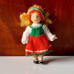 May include: A porcelain doll with blonde hair wearing a red, green, and white knitted dress and a red headband.