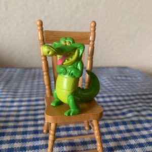 May include: A green and yellow alligator figurine with a pink tongue, sitting on a small wooden chair. The alligator has a smiling expression. The chair is made of light brown wood.