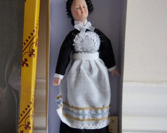 May include: A porcelain doll wearing a black dress with a white apron and a white lace collar. The doll has black hair and brown eyes. The doll is packaged in a white box with a small label.