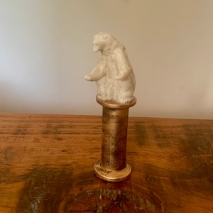 Puede incluir: Una figurita de oso de cerámica blanca de pie sobre un pedestal de metal dorado. El oso está mirando hacia la izquierda y tiene un diseño simple.