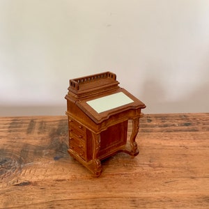 Puede incluir: Un escritorio de madera en miniatura con una tapa inclinada y un cajón. El escritorio está hecho de madera oscura y tiene una tapa marrón claro.