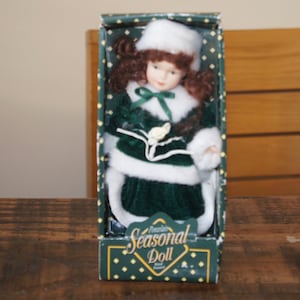 May include: A porcelain doll in a green velvet dress with white fur trim and a white hat. The doll is packaged in a box with gold lettering that reads "Seasonal Doll".
