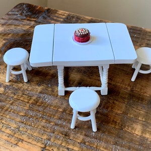 Puede incluir: Una mesa blanca con un pastel blanco en un plato, rodeada de cuatro taburetes blancos. La mesa y los taburetes están sobre una superficie de madera.