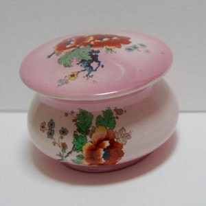 May include: A small, pink ceramic jar with a lid. The jar and lid are decorated with a floral pattern in shades of orange, red, green, and yellow.