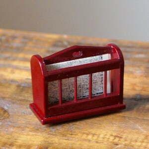 May include: A small, red wooden magazine rack with a decorative top and a white paper insert. The rack is sitting on a wooden surface.