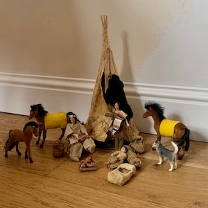 May include: A Native American themed playset featuring a teepee, two figures, two horses, a dog, and a baby doll. The teepee is made of brown fabric with a yellow and brown pattern. The figures are dressed in traditional clothing. The horses are brown with yellow blankets. The dog is grey and white. The baby doll is wearing a brown blanket.