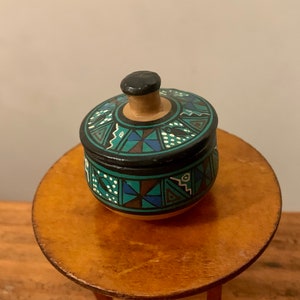 Pode incluir: Um pequeno recipiente redondo de cerâmica com tampa. O recipiente é decorado com um padrão geométrico em tons de preto, verde, azul e branco.