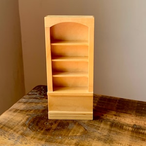 Op de afbeelding: Een miniatuur houten boekenkast met vier planken en een gewelfde bovenkant. De kleine kast is gemaakt van lichtgekleurd hout en staat op een houten oppervlak. De boekenkast is een decoratief item.