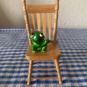 Op de afbeelding: Een kleine houten stoel met een groene kikkerfiguur. De kikker heeft grote ogen en houdt een klein voorwerp vast. De stoel is lichtbruin en heeft een klassiek ontwerp. De achtergrond is een blauw-wit geruit patroon.