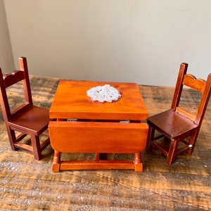 May include: A miniature wooden table set with two chairs. The table has a drop leaf and is made of a light brown wood. The chairs are also made of light brown wood and have a simple design.