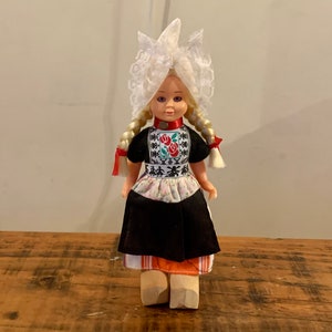 Könnte beinhalten: Eine kleine Puppe in traditioneller niederländischer Tracht. Die Puppe hat blonde Haare, blaue Augen und trägt eine weiße Spitzenhaube, ein schwarzes Kleid mit einer weißen Schürze und orange-weiße Holzschuhe.