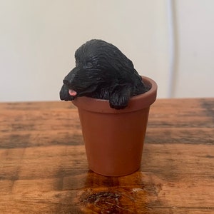 Pode incluir: Uma figura de cão preto está sentada dentro de um vaso de cerâmica marrom. O cão está olhando para a esquerda e tem a língua de fora.