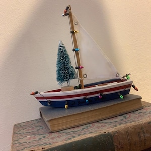 Puede incluir: Un velero decorativo de madera con vela blanca, casco rojo y azul, y un pequeño árbol de Navidad. El barco está adornado con luces de colores y descansa sobre dos libros antiguos. El barco tiene el texto "St. Joe" en el lateral.