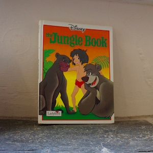 Puede incluir: Un libro infantil vintage de Disney El Libro de la Selva con una portada roja, naranja y verde. La portada presenta a Mowgli, Baloo y Bagheera. El libro est&aacute; publicado por Ladybird.