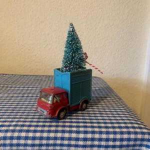 May include: A red toy truck with a blue box bed carrying a small green artificial Christmas tree with white flocking. The truck is on a blue and white checkered tablecloth.