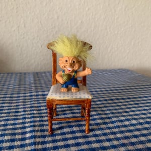 Puede incluir: Un muñeco troll de pelo amarillo con un mono azul y un micrófono en la mano, sentado en una silla de madera con un asiento a cuadros azules y blancos.