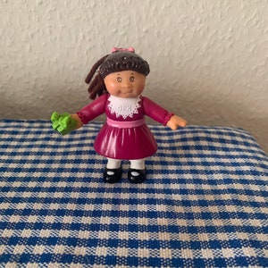 Könnte beinhalten: Eine kleine Plastikpuppe mit braunen Haaren, die ein burgunderfarbenes Kleid mit einem weißen Spitzenkragen und einer rosa Schleife trägt. Die Puppe hält ein grünes Objekt in ihrer linken Hand und hat ein weißes Lätzchen mit dem Text "CPK" darauf.