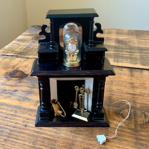 May include: A miniature black wooden fireplace mantel with a gold clock under a glass dome. The fireplace has a white interior and includes a set of gold fireplace tools.