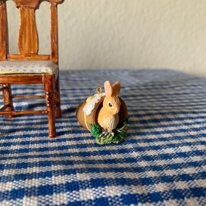 Pode incluir: Uma pequena figura de coelho de cerâmica marrom está espiando de uma cesta com folhas verdes. A figura está sentada em uma toalha de mesa quadriculada azul e branca.
