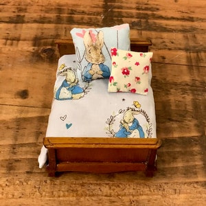 Puede incluir: Una cama de madera en miniatura con un juego de ropa de cama azul y blanco con ilustraciones de Peter Rabbit. La cama tiene una almohada blanca con flores rojas y una almohada azul con una ilustración de Peter Rabbit.