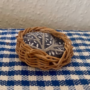 以下が含まれることがあります： グレーと白のパターンが施されたファブリックのクッションが入った、小さく丸い籐製のバスケット