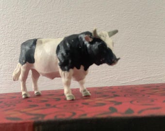 May include: A small, decorative figurine of a bull. The bull has a white body with black patches on its back and head. It has a single horn and is standing on a red patterned surface.