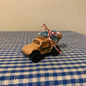 Puede incluir: Un pequeño coche de juguete de color beige con un pequeño árbol de Navidad verde flocado en la parte superior. El coche tiene ruedas negras y una cuerda roja y blanca atada alrededor del árbol. El coche está sentado sobre un mantel a cuadros azul y blanco.