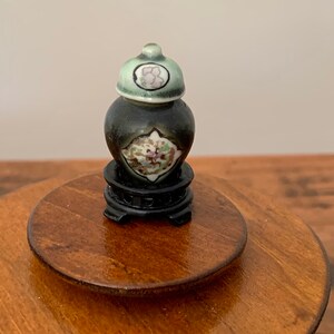 Könnte beinhalten: Eine kleine schwarze und grüne Keramikvase mit Deckel und Blumenmuster, die auf einem Holzsockel steht.