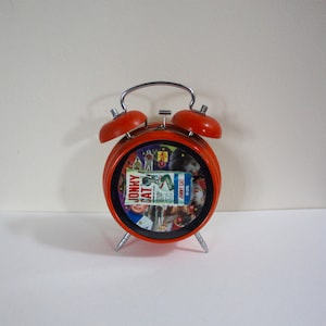 May include: Orange alarm clock with a black face and a colorful image of a magazine cover with the text "Jonny Cat" on it.