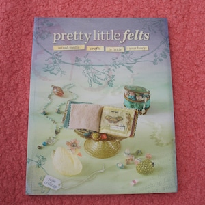 May include: A book titled "pretty little felts" with the subtitle "mixed-media crafts to tickle your fancy". The book cover features a small, handmade book with a butterfly design, a white porcelain duck, and a variety of colorful beads and trinkets.