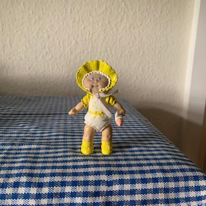 May include: A yellow Cabbage Patch Kid doll wearing a yellow bonnet and a white dress with yellow flowers. The doll has blue eyes and a bandage on its arm.
