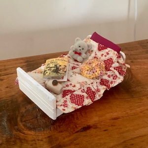 May include: A miniature white bed with a red and white floral quilt, a yellow book, and a gray teddy bear with a red bow.