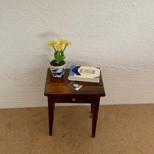 May include: A small wooden table with a drawer, a book, a photograph, and a potted plant with yellow flowers. The table is brown and the flowers are yellow.