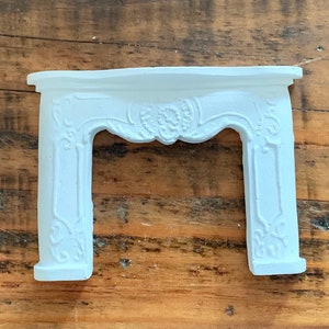 May include: White painted miniature fireplace mantel with ornate detailing. The mantel is made of a lightweight material and has a simple, elegant design.