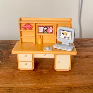 Puede incluir: Un escritorio de madera en miniatura con una computadora, un ratón y una estantería con un coche de juguete rojo, lápices y un tablero de anuncios con papeles.