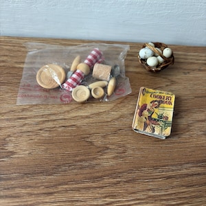 Puede incluir: Accesorios de cocina en miniatura que incluyen un cuenco de madera pequeño, un elemento a cuadros rojos y blancos y un pequeño libro de cocina. Una pequeña cesta contiene varios huevos blancos. El libro de cocina se titula "Round-the-Clock Cookery Book."