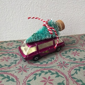 Può includere: Un'auto giocattolo rosa con un piccolo albero di Natale verde legato al tetto con spago rosso e bianco. L'auto ha un tappo di sughero di legno sopra l'albero. L'auto giocattolo è su una superficie a motivi.