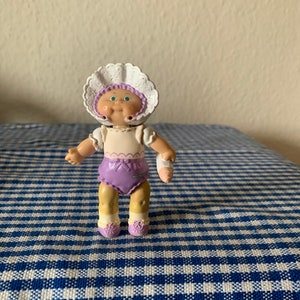 Pode incluir: Uma pequena boneca Cabbage Patch Kid de plástico vestindo um gorro branco com um vestido branco e roxo e meias brancas. A boneca tem olhos verdes e rosto corado.