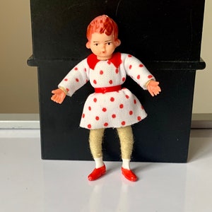 May include: A small plastic doll with red hair, wearing a white dress with red polka dots, a red collar, and red shoes.